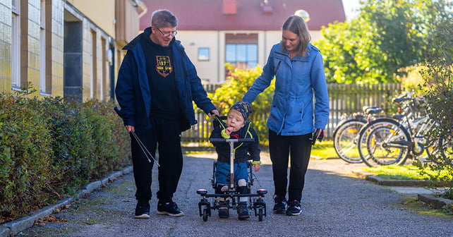 Ung gutt går i sin NF-Walker og holder foreldrenes hånd.