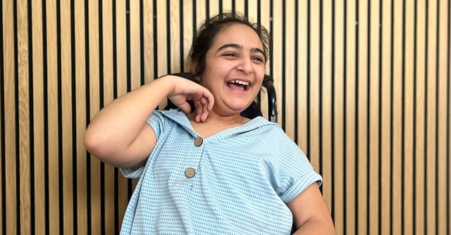 Alma, en smilende jente med CP, sitter foran en vegg med trespiler og ler med hånden løftet ved halsen.