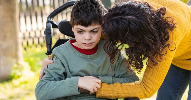 Mother hugging her son with disability sitting in a wheelchair in a park, showing love and support.