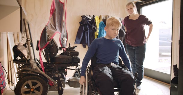 A boy with Duchenne muscular dystrophy sits in a wheelchair at home, with his mother standing in the background and assistive mobility equipment nearby.