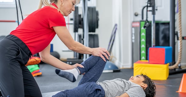 En kvinnlig fysioterapeut arbetar med en ung pojke i ett gym när hon stretchar och stärker hans ben.