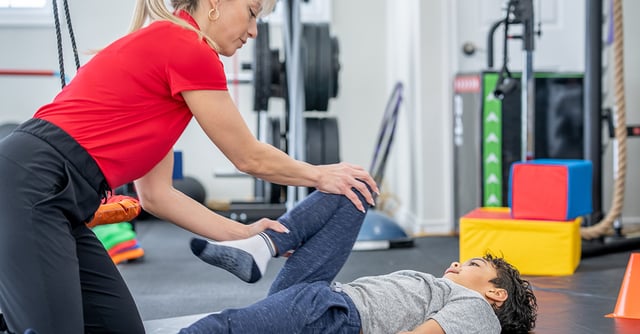 Eine Physiotherapeutin arbeitet mit einem jungen Jungen in einem Fitnessraum, während sie seine Beine dehnt und stärkt.