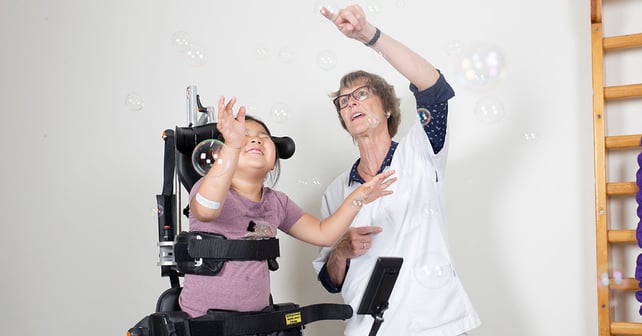 A young girl stands in Innowalk while blowing soap bubbles with her physiotherapist.