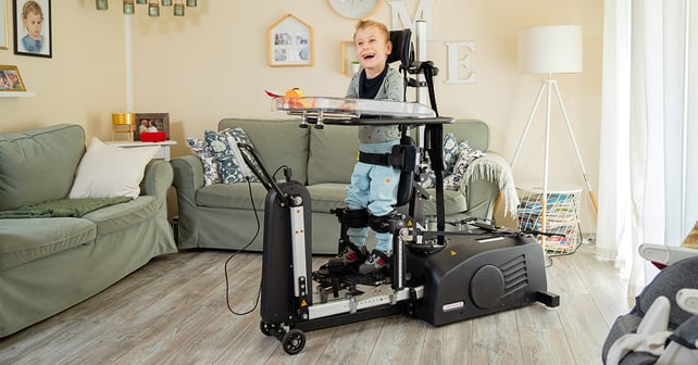 A smiling boy uses an Innowalk a dynamic standing device in a living room with sofas and family photos in the background.