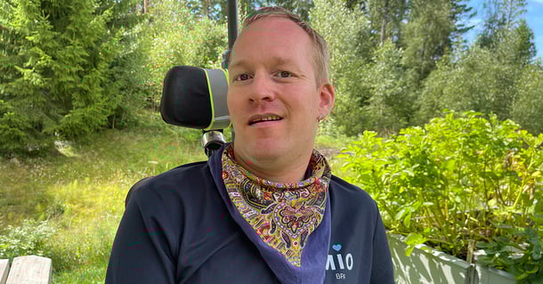 Ein Mann sitzt im Rollstuhl im Freien, lächelt und trägt ein buntes Halstuch, mit Bäumen im Hintergrund.
