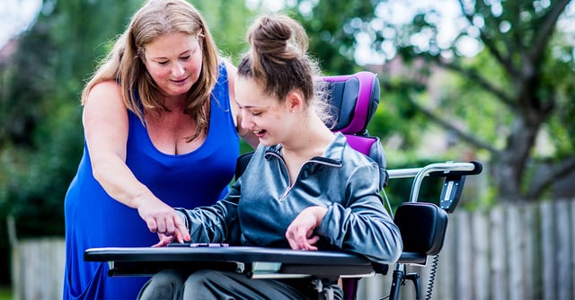 Kvinne hjelper en jente i en BeMe sittemodul med aktivitet ved bord, utendørs i grønne omgivelser.