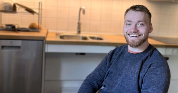 Leende person med kort skägg som sitter i ett kök och bär en mörk tröja.