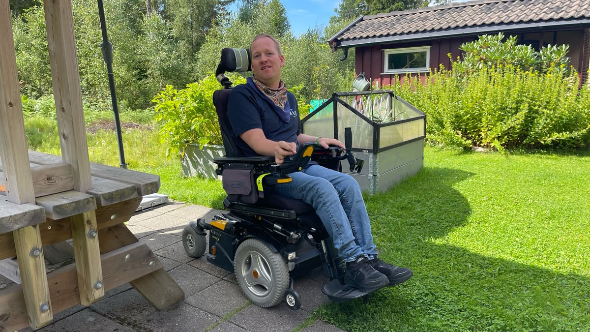 Mann im elektrischen Rollstuhl im Freien neben einem Haus, lächelnd in der Sonne.