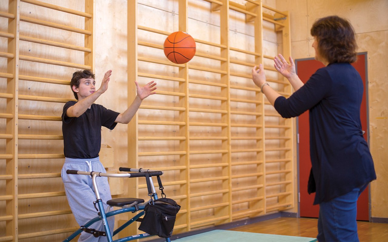 Junger Mann mit Rollator spielt Ball mit einer Frau vor einer Sprossenwand.