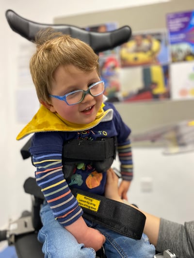 A smiling young boy wearing glasses is supported in an Innowalk device, leaning slightly forward while an adult assists him in a school setting.